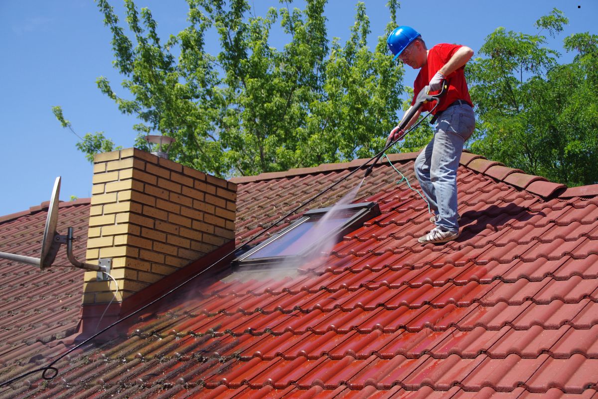 A man in safety gear uses a pressure washer to clean a roof, ensuring a thorough and safe cleaning process.