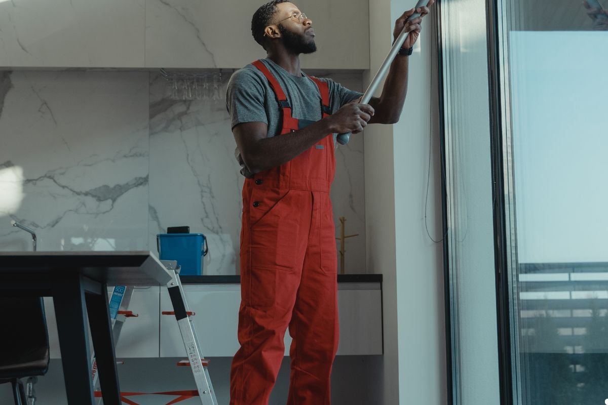 A man in overalls holds a ladder while painting a window, showcasing a home improvement project in progress. 